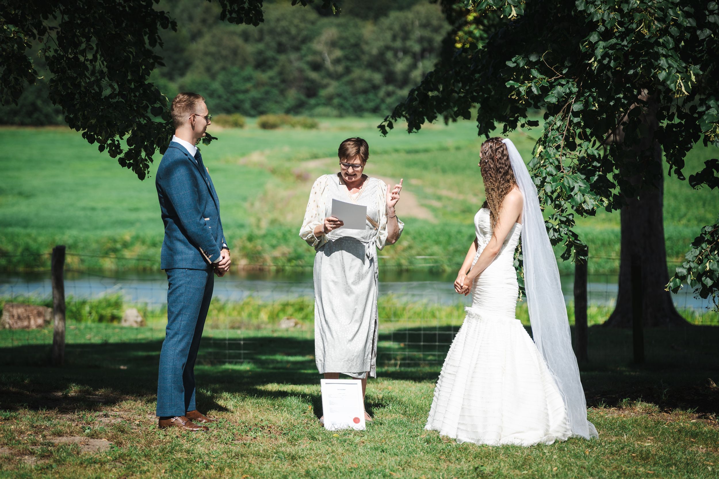 A wedding party walks together on lush green grass near a lake on a sunny summer afternoon.