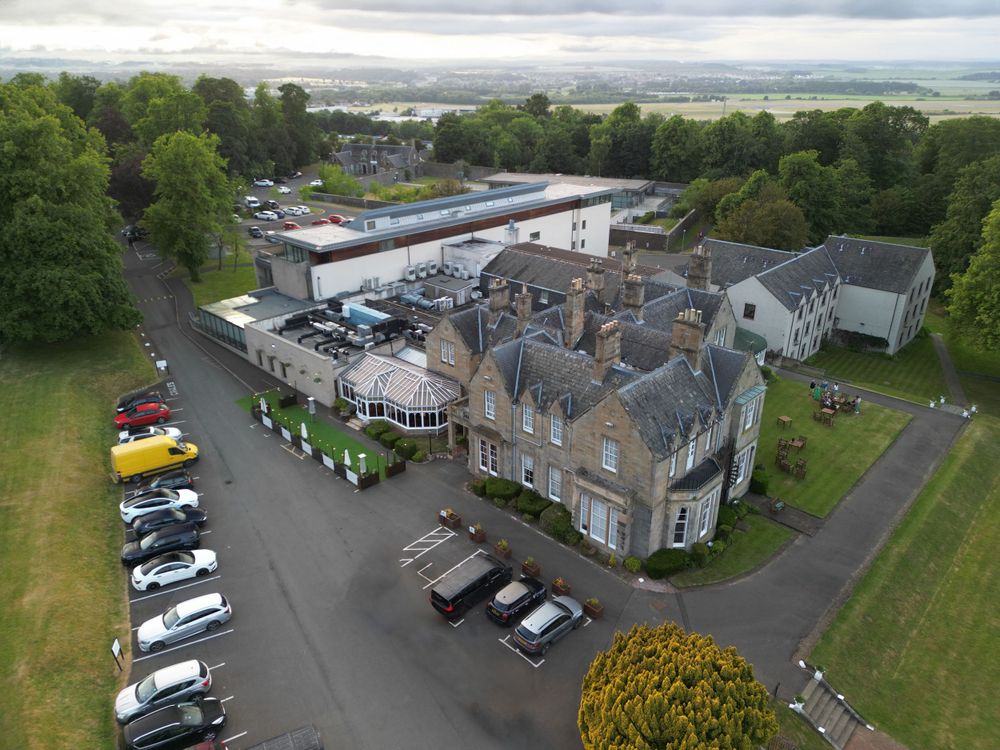 Aerial view of a historic stone mansion with parking lot and surrounding gardens in Scotland.