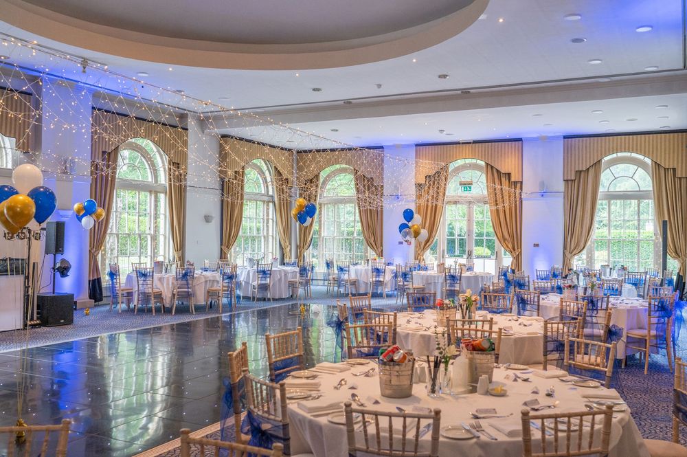 Elegant ballroom with circular tables, arched windows, and blue uplighting reflecting on polished marble floors.