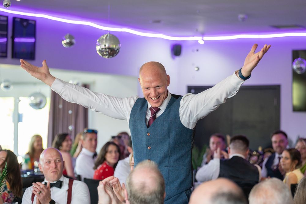 Wedding reception guests celebrating and dancing under purple mood lighting in an indoor venue.