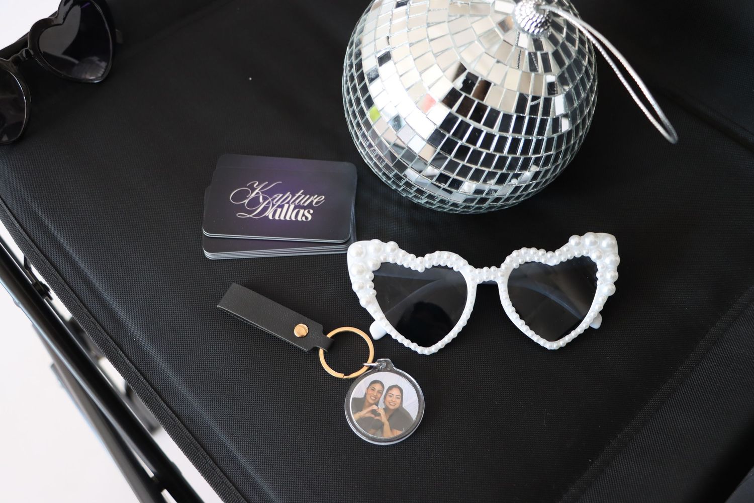 White pearl heart sunglasses, disco ball, keychain photo, and Kristure Dallas cards on black surface.