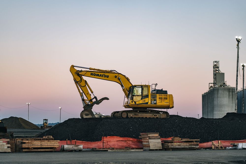 Heavy machinery and excavators operate at an industrial site during sunset with silhouettes against the orange sky.