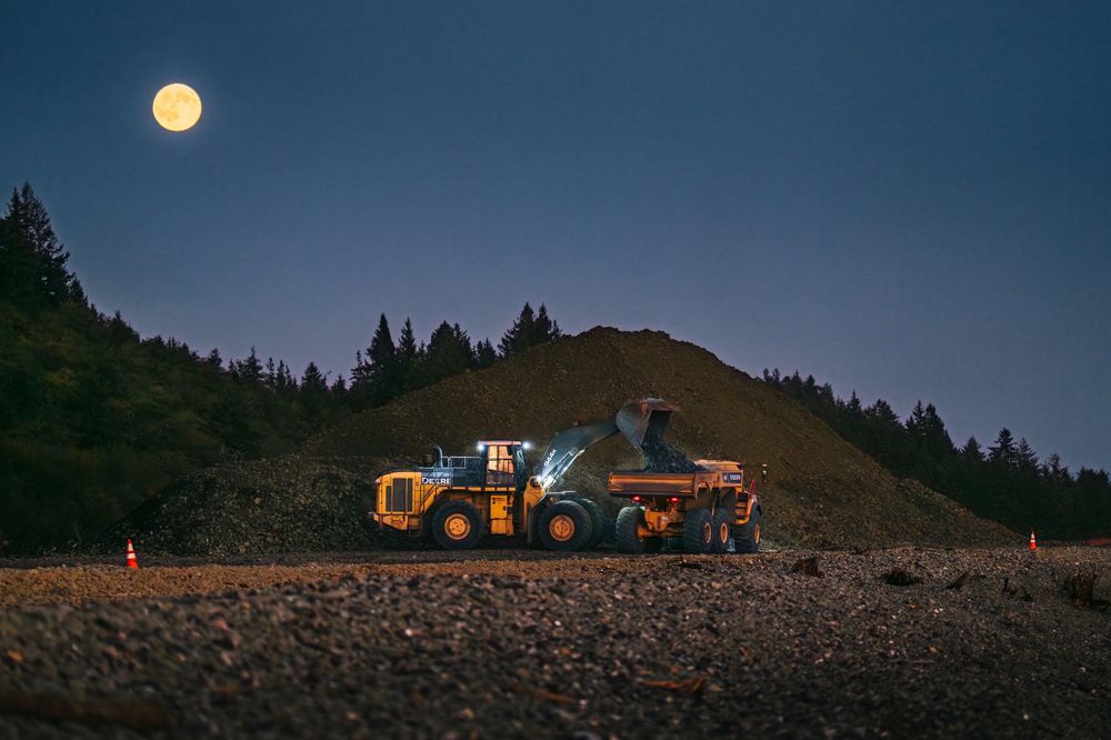 Heavy machinery silhouetted against a moonlit sky on a construction site with trees in the background.