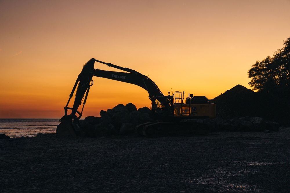 Construction equipment silhouettes against a vibrant orange sunset sky at a coastal worksite.
