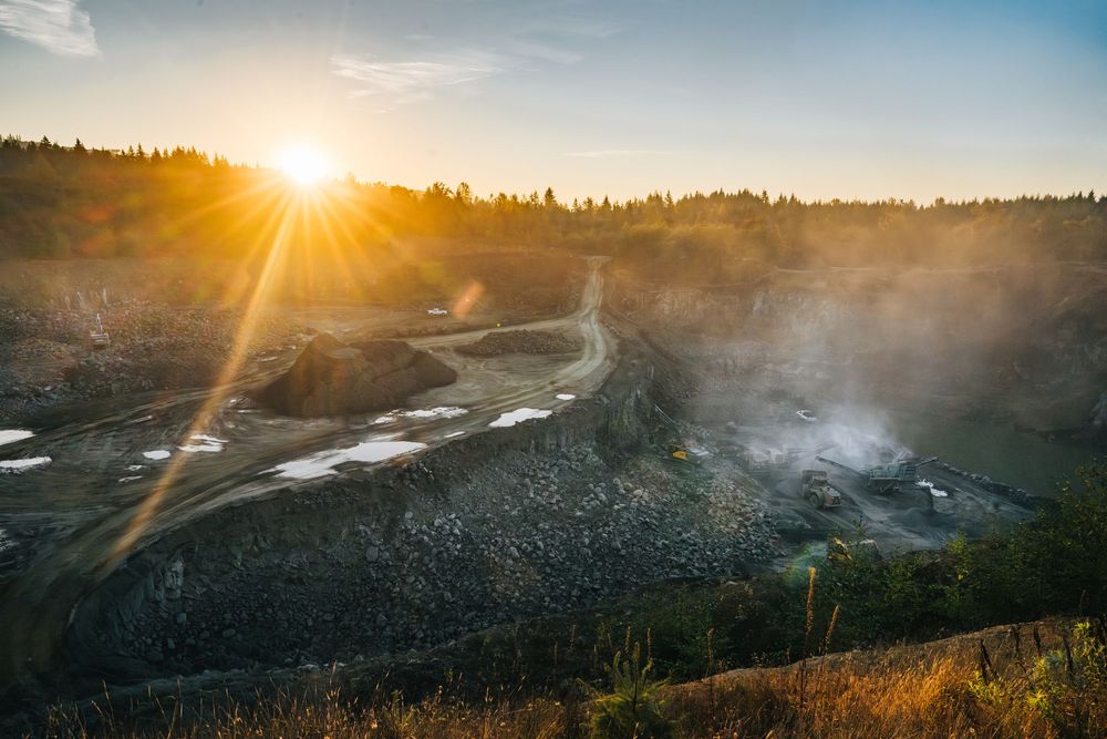 Construction equipment working at sunset on a rocky terrain with sunburst and mountain landscape.