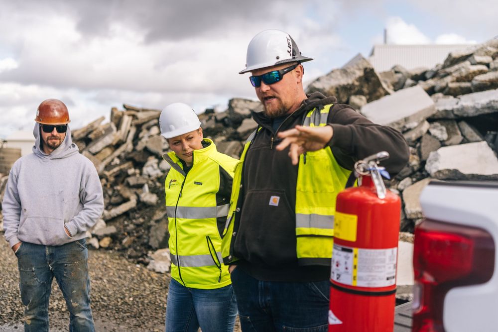 Construction workers in safety vests inspect equipment and discuss plans at a rocky worksite.