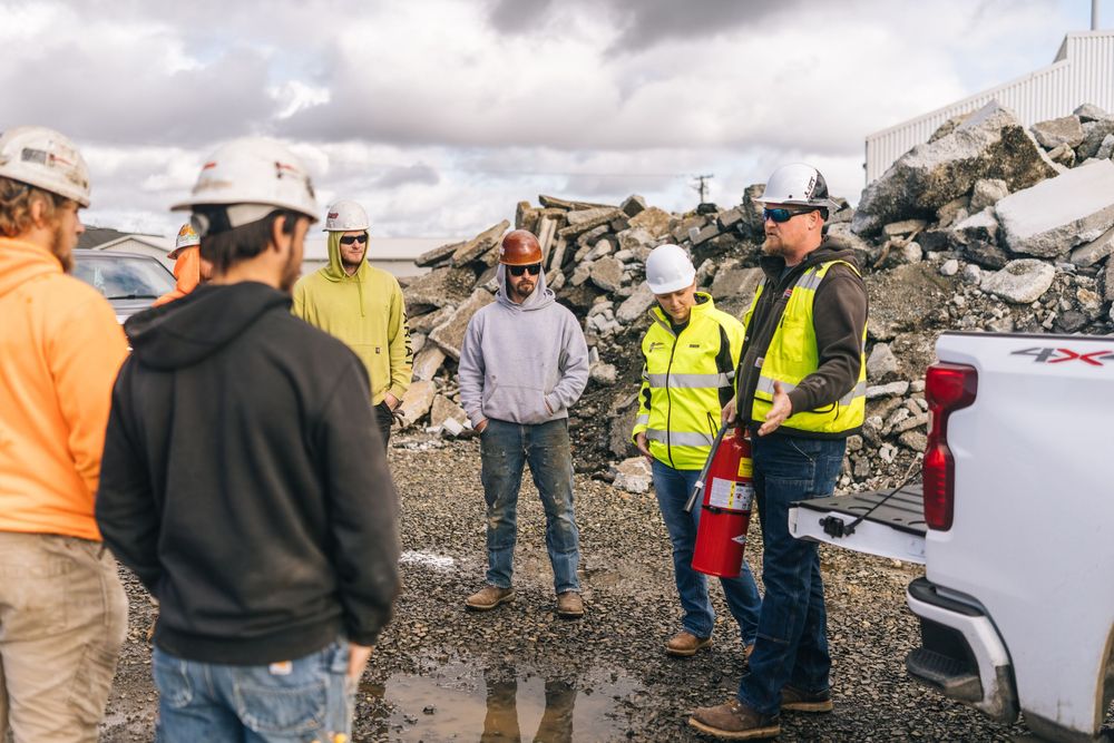 Workers in safety gear and hard hats gather at a construction or demolition site with debris piles visible in background.
