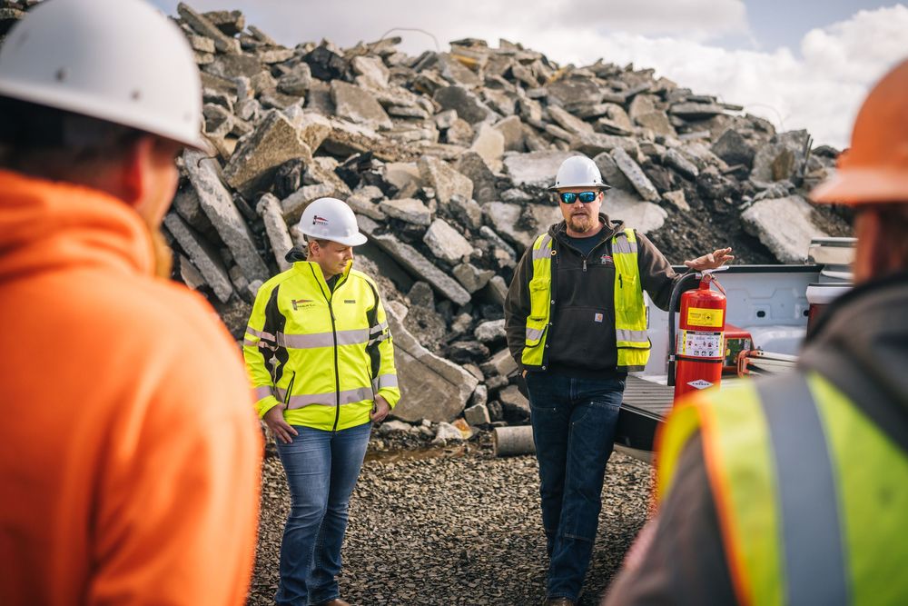 Workers in high visibility safety vests gather at a construction or demolition site with large piles of debris and rubble.