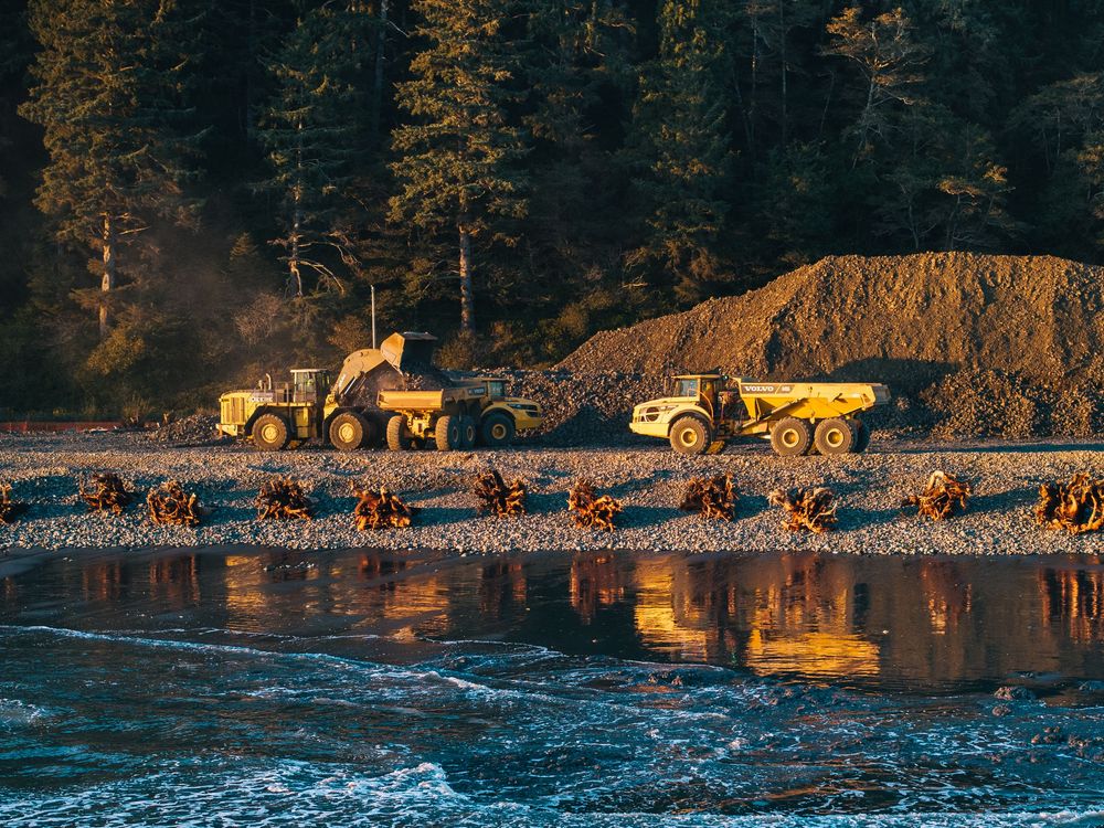 Heavy construction dump trucks and loader working near a reflective water's edge at sunset with evergreen forest backdrop.