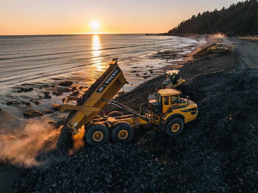 Mining trucks and excavators work along a rocky coastline at sunset with golden light reflecting on the water.