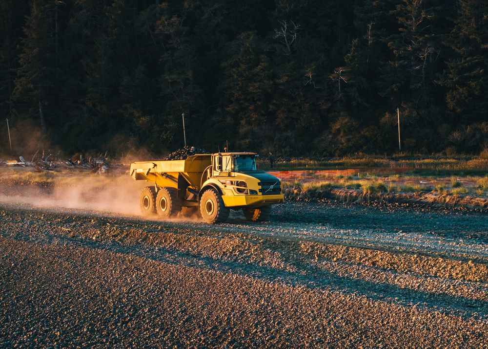 Yellow construction equipment working on a gravel surface at dusk with dark forest in the background.