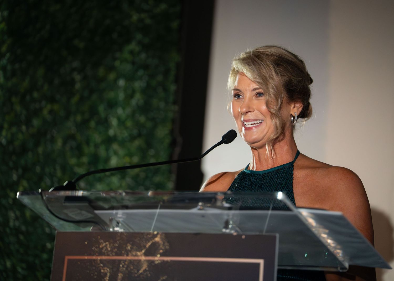 A speaker smiles while delivering remarks at a podium with a green hedge backdrop.