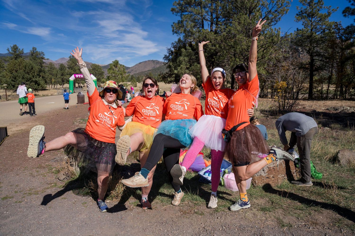 A group of people in orange shirts pose playfully on the ground against a backdrop of pine trees and blue sky.