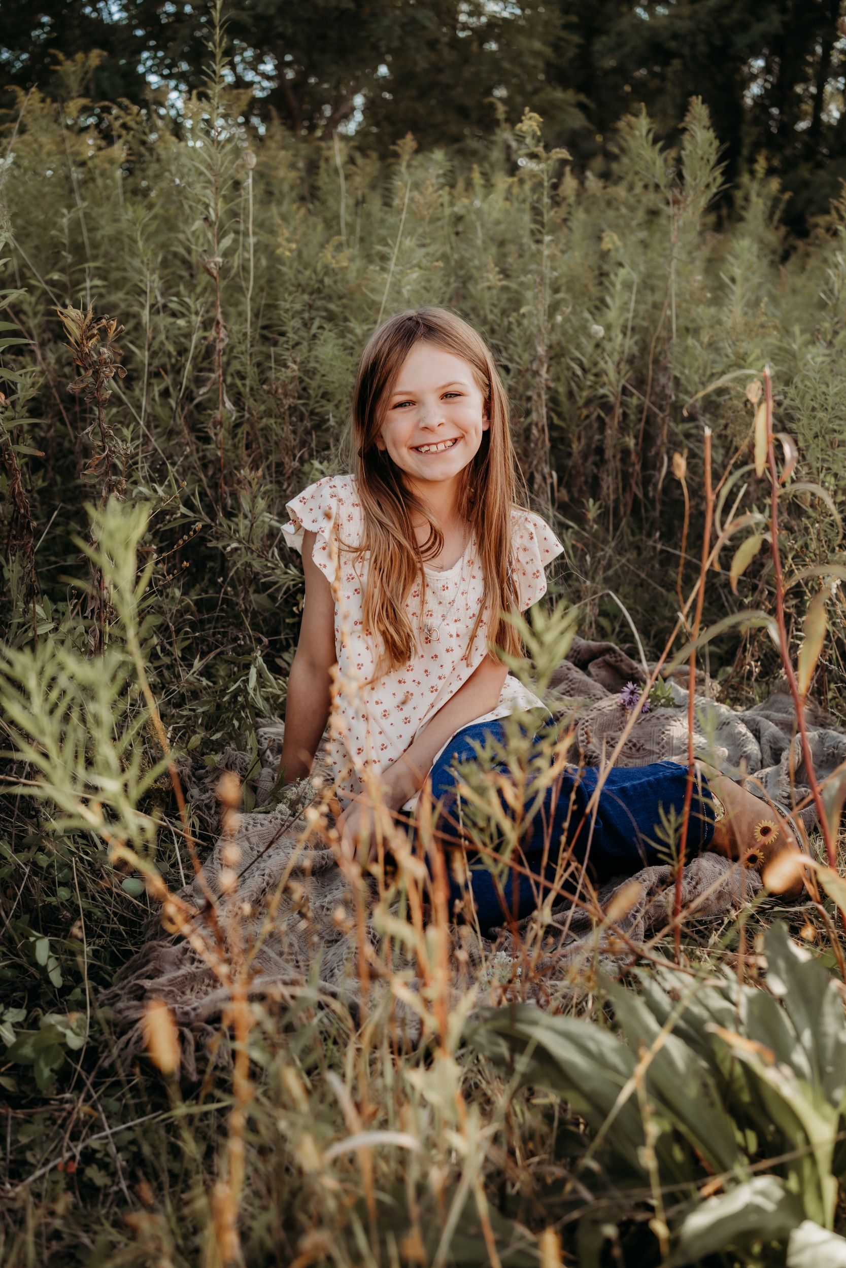 Young child playing and smiling while sitting in a field of tall grass and wildflowers.