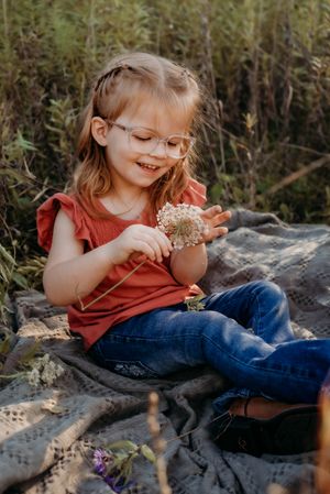 A child in a rust-colored shirt and jeans sits on a rocky surface in a natural outdoor setting with greenery in background.