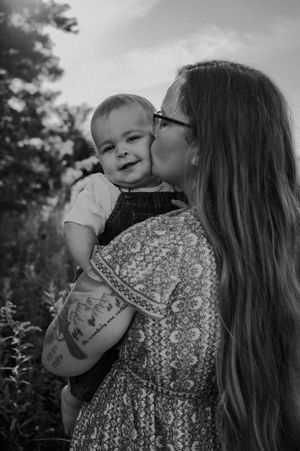 A tender black and white portrait of a mother in a patterned dress kissing her baby wearing overalls outdoors.