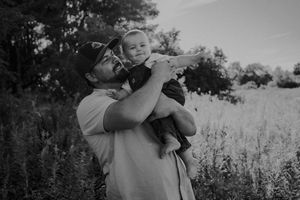 Black and white photo of couple embracing in a field with wildflowers and trees in the background.