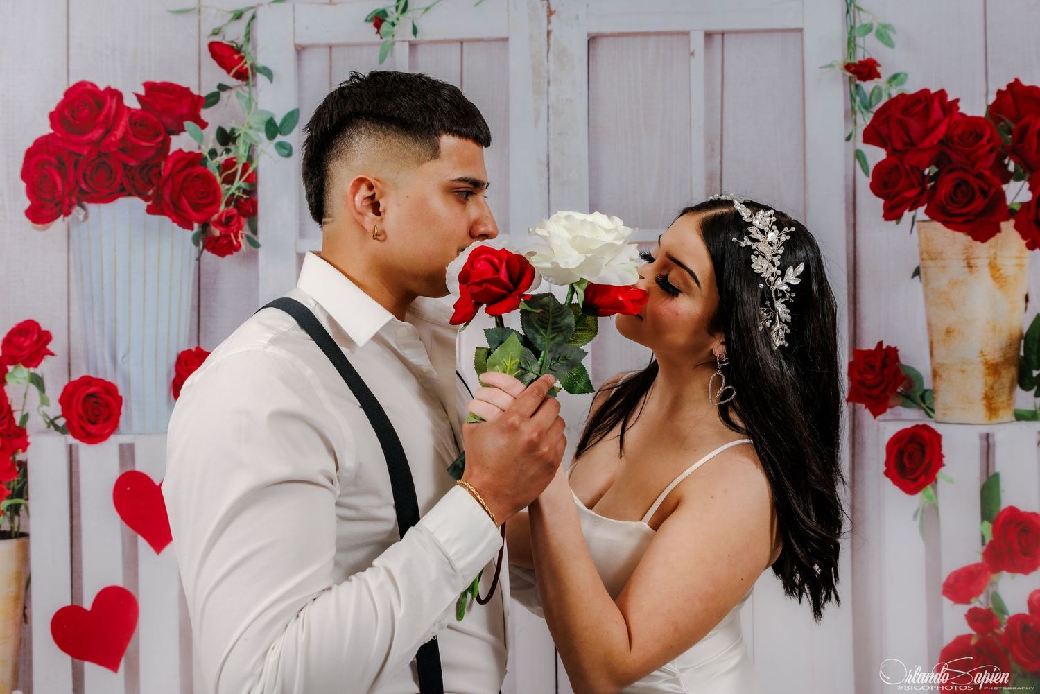 A romantic couple shares a tender moment surrounded by red roses and white decor in a wedding setting.