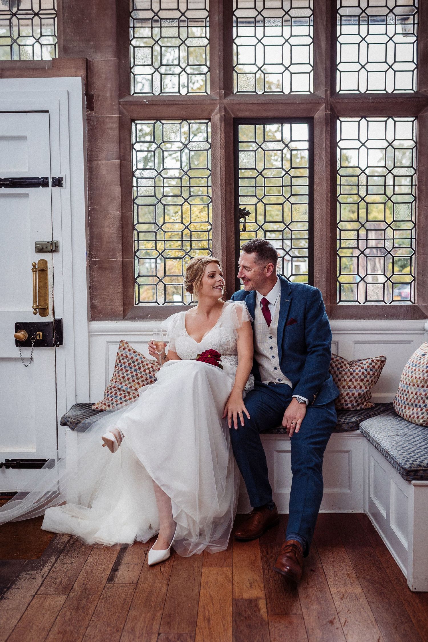 Newlywed couple shares a joyful moment sitting together on a window bench with ornate glass panels and wood floors.