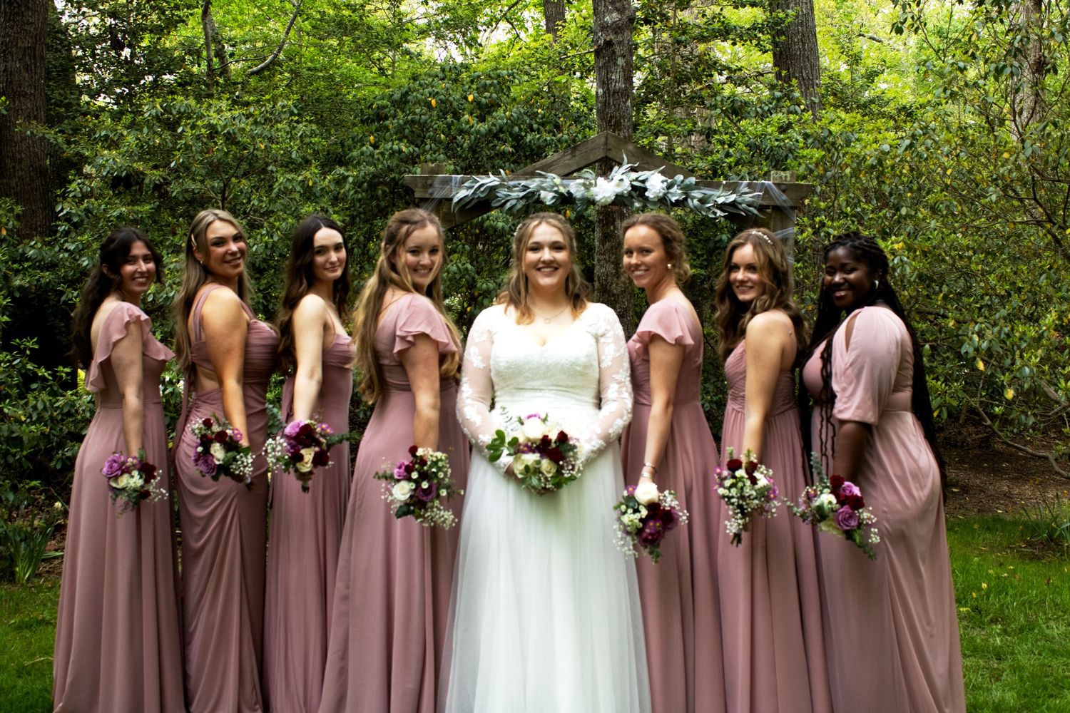 Bridal party wearing mauve dresses poses with bride in white gown outdoors.