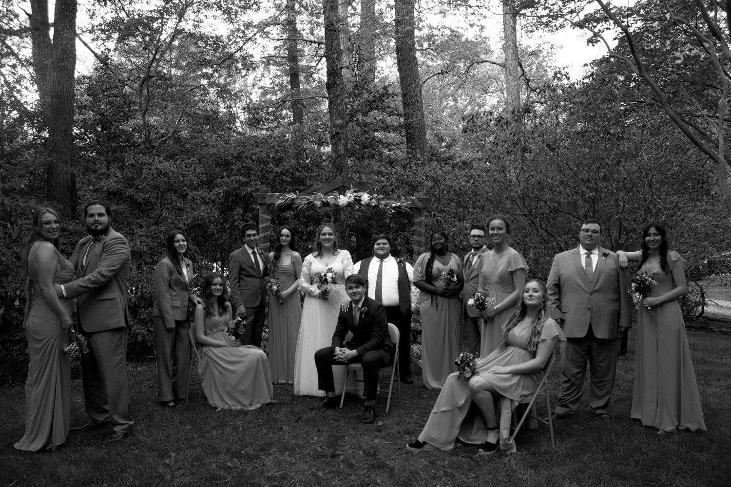 Large wedding party gathered for group photos in a wooded outdoor setting.