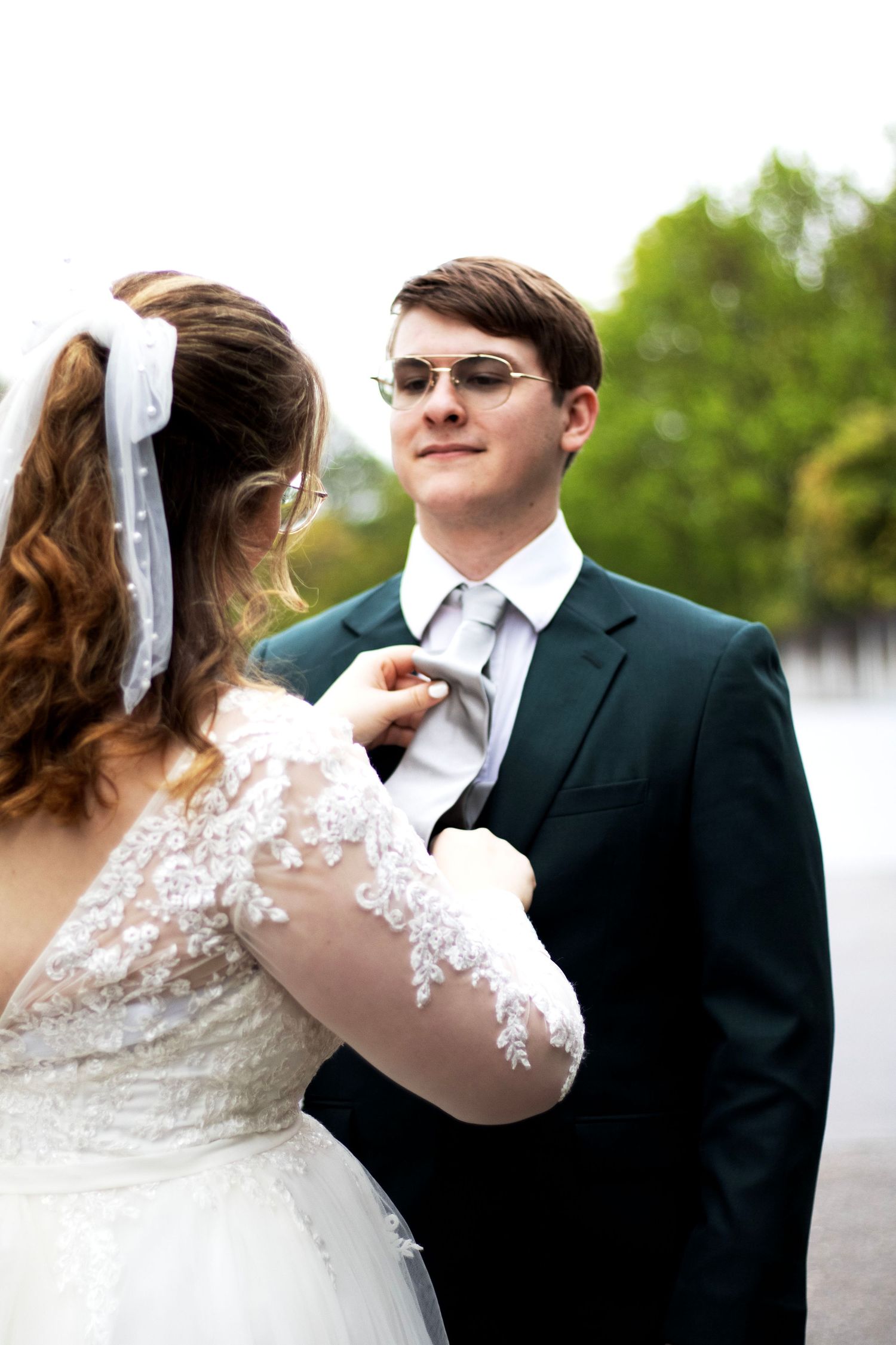 Elegant wedding couple in formal attire with lace details and sunglasses outdoors.