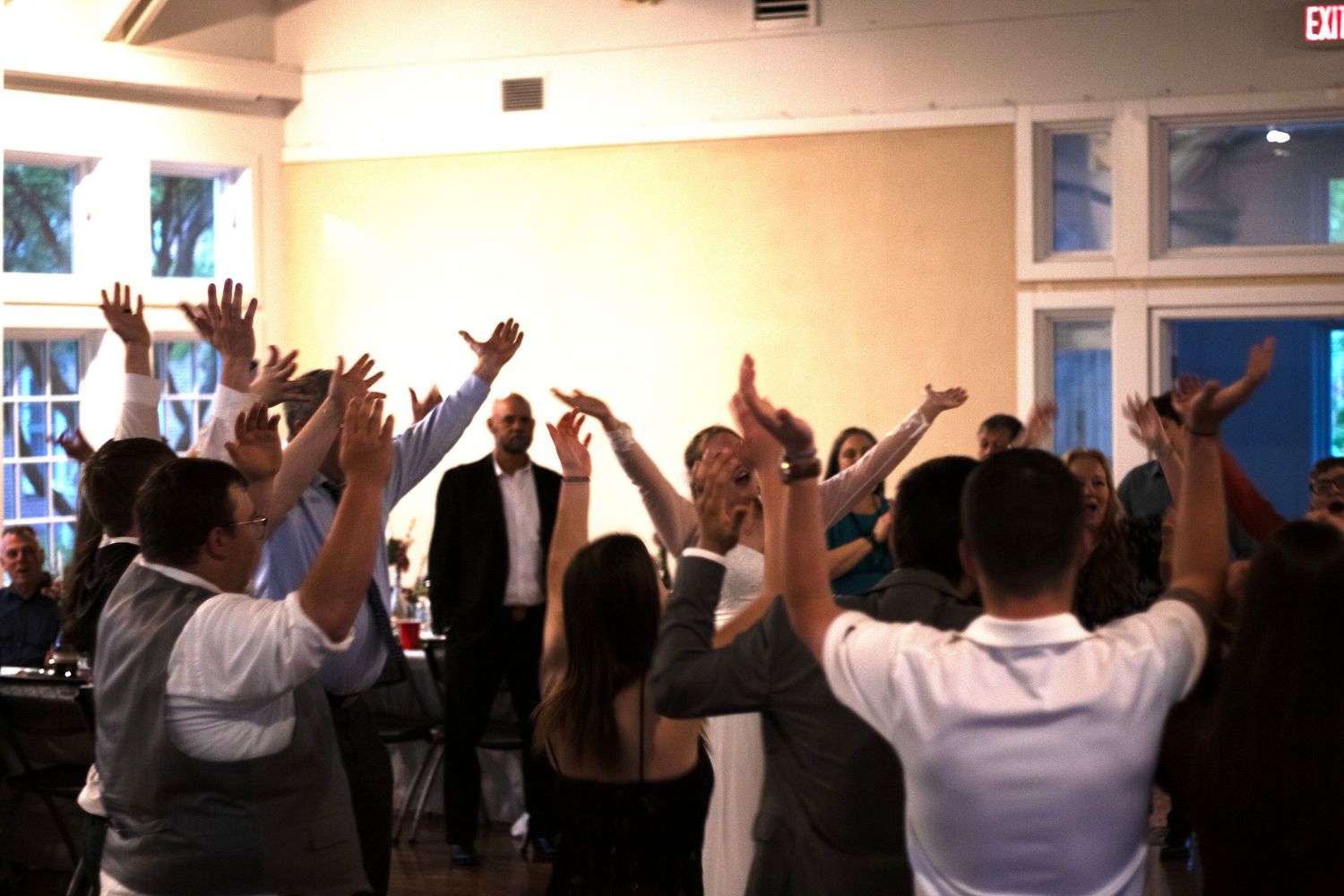 Wedding reception crowd with raised hands celebrating in an indoor venue.