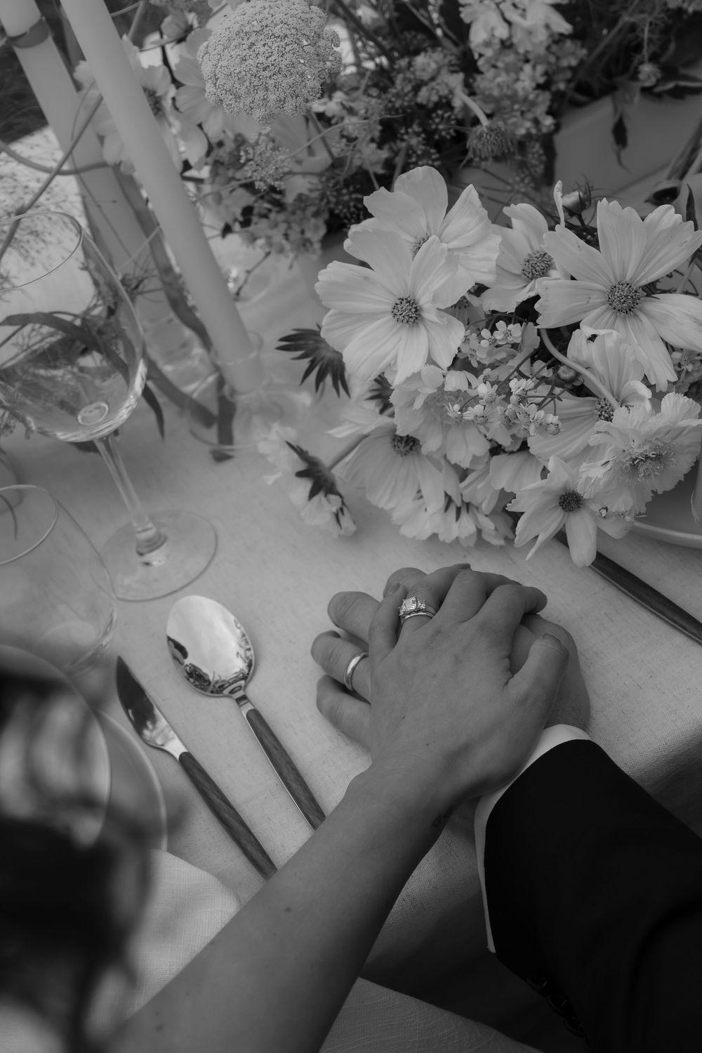A black and white photo showing hands reaching towards delicate flowers and petals arranged on a surface.