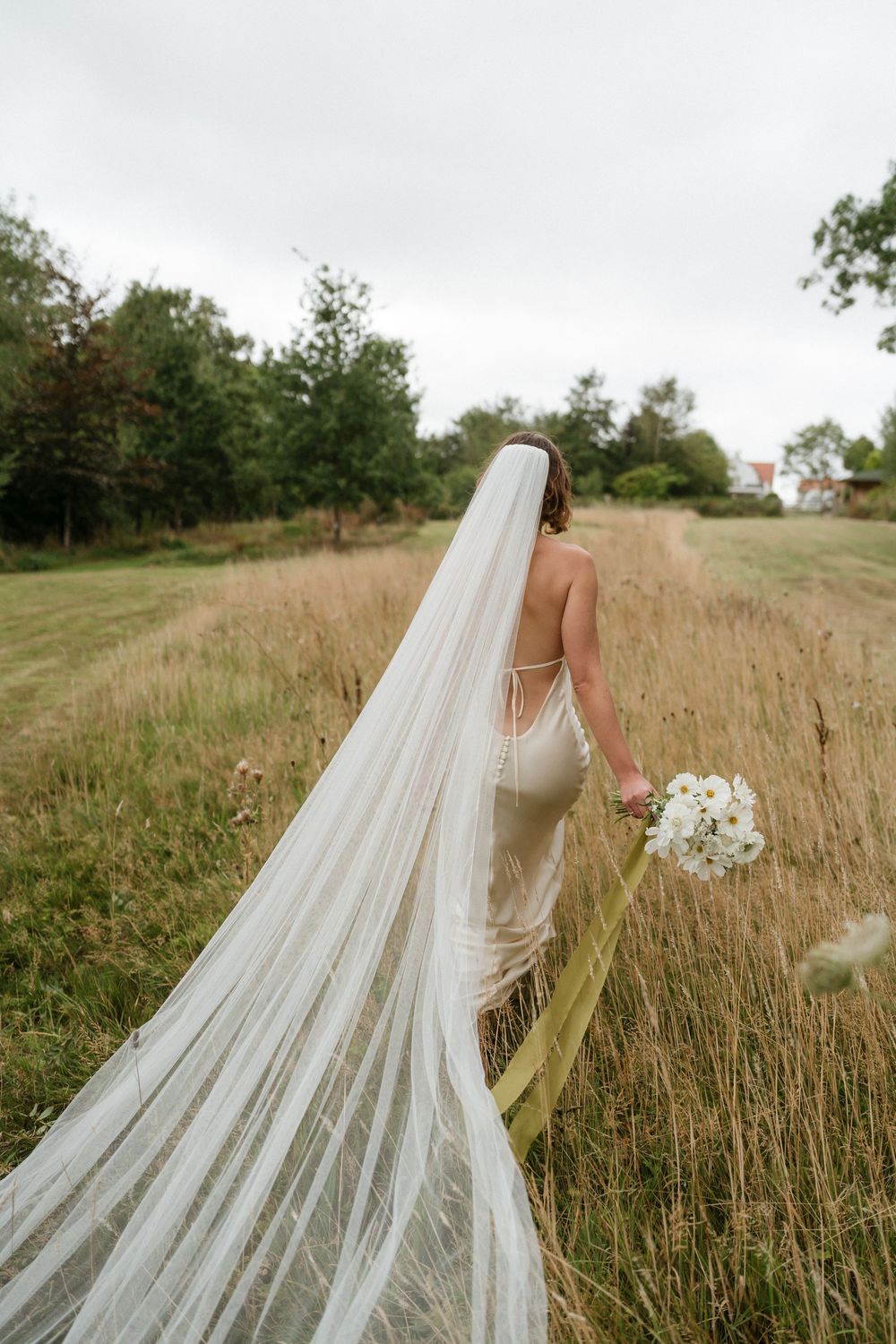A flowing cathedral-length veil trails behind a bride in a white gown as she walks through a golden wheat field.