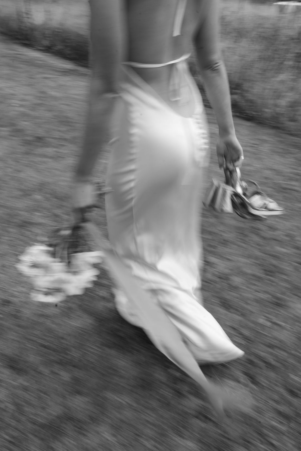 A blurred black and white photo of a bride walking on grass, holding her shoes and bouquet.