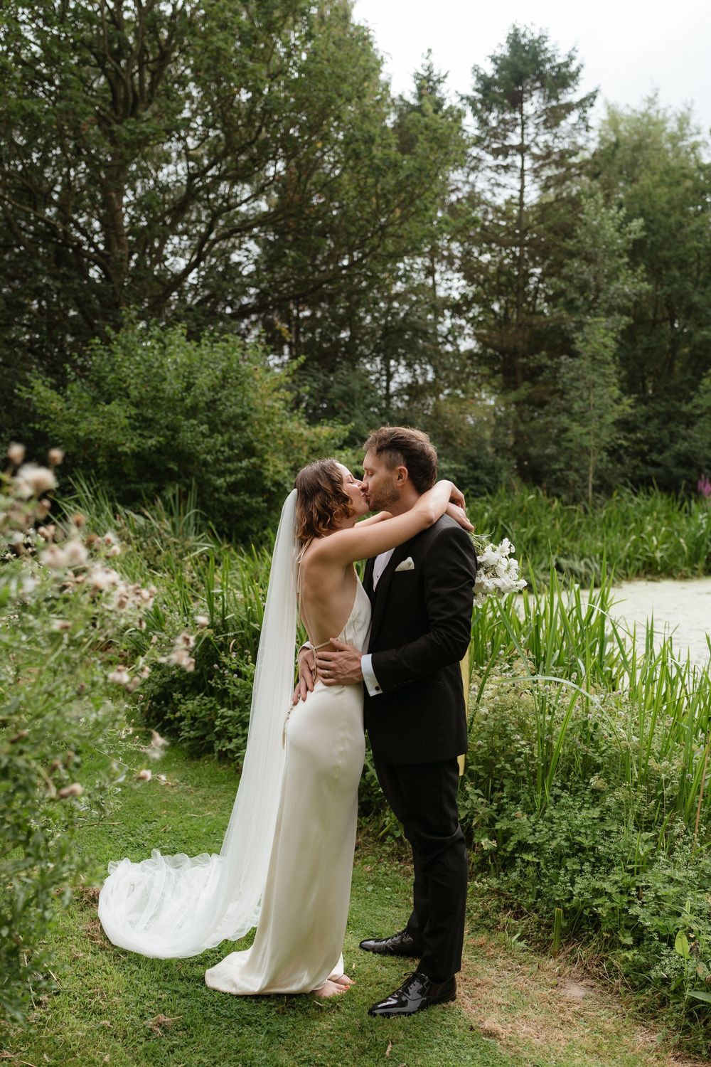 A couple shares a romantic kiss on their wedding day in a lush garden setting with evergreen trees in the background.