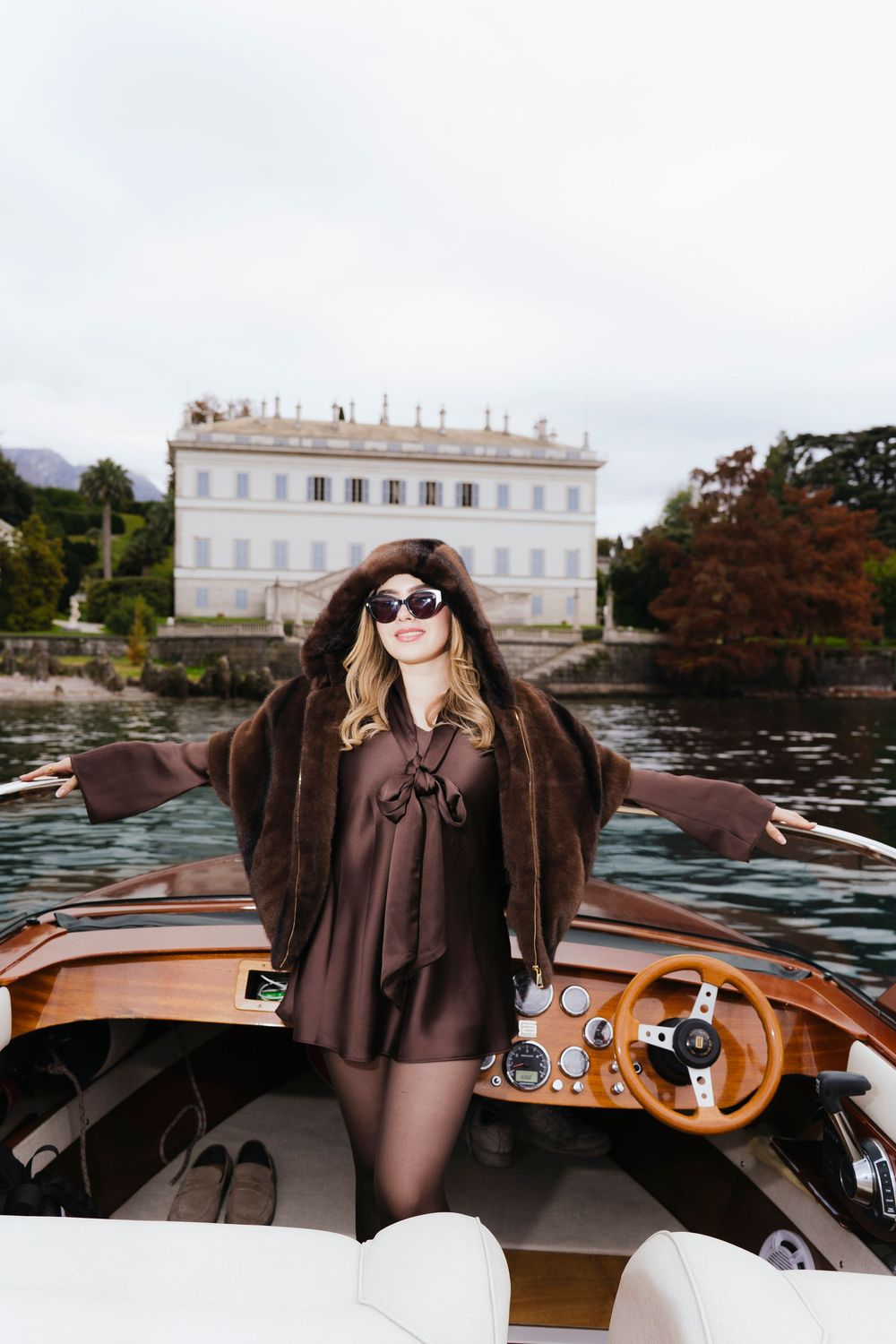 A person wearing sunglasses and a brown dress sits in a vintage wooden boat on a scenic lake.