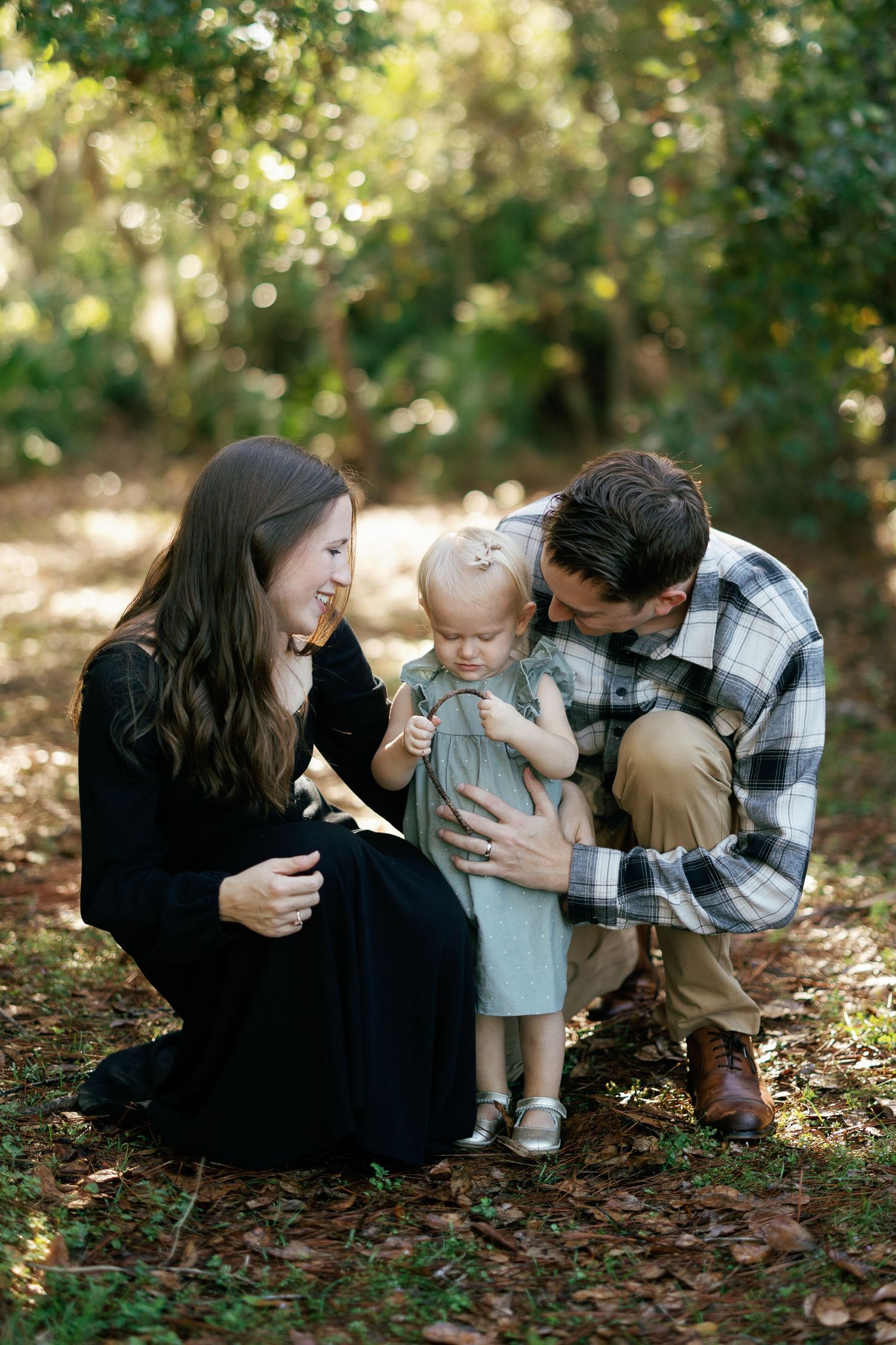 A family in casual fall attire sits together on fallen autumn leaves in a sun-dappled forest setting.