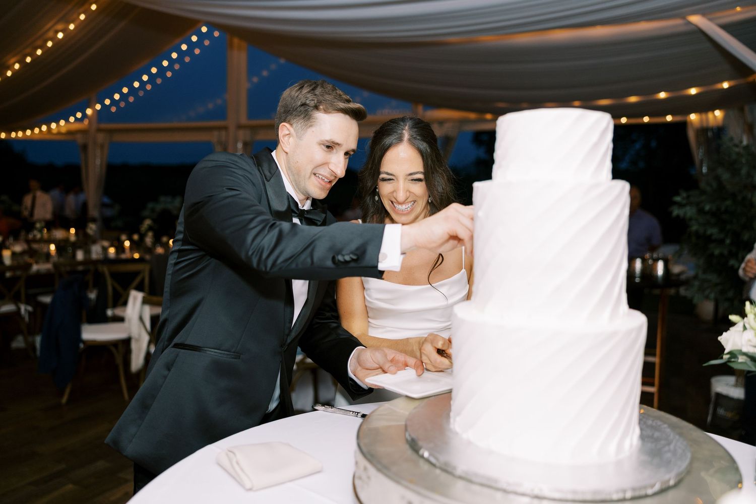 A couple stands together at a wedding reception with a white tiered cake in an outdoor venue with string lights overhead.