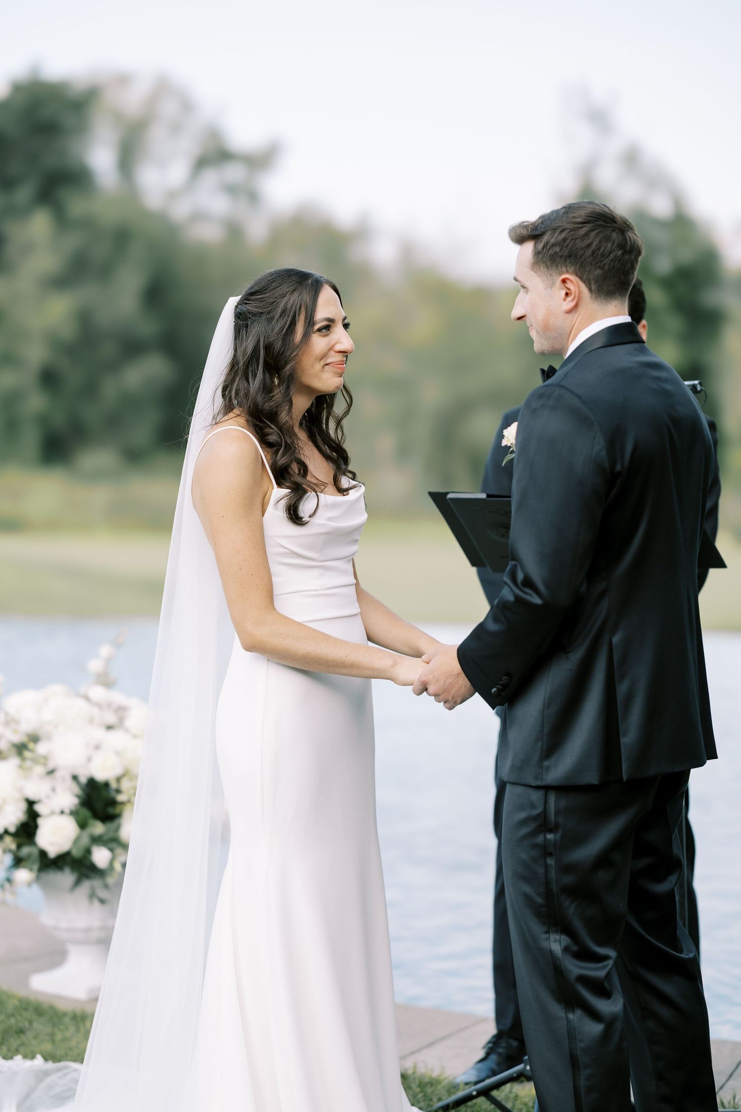 A couple exchanges vows during an outdoor wedding ceremony by a lake with white floral decorations.
