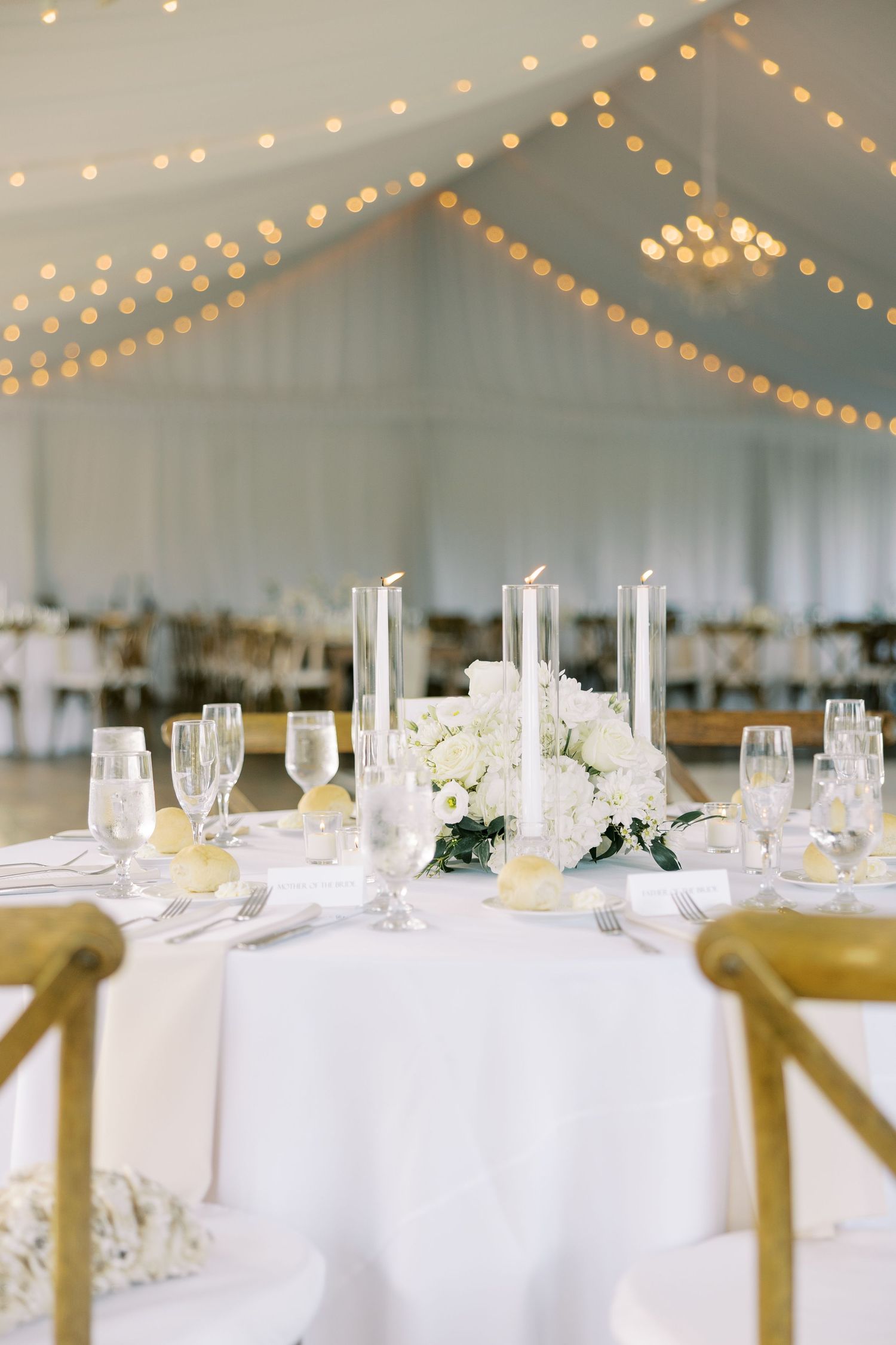 Elegant wedding reception tables with white linens, string lights above, and delicate floral centerpieces in a tent setting.