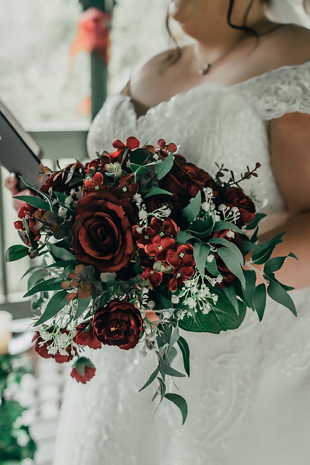 Dark burgundy roses with green eucalyptus leaves create a dramatic bridal bouquet against a white wedding dress background.