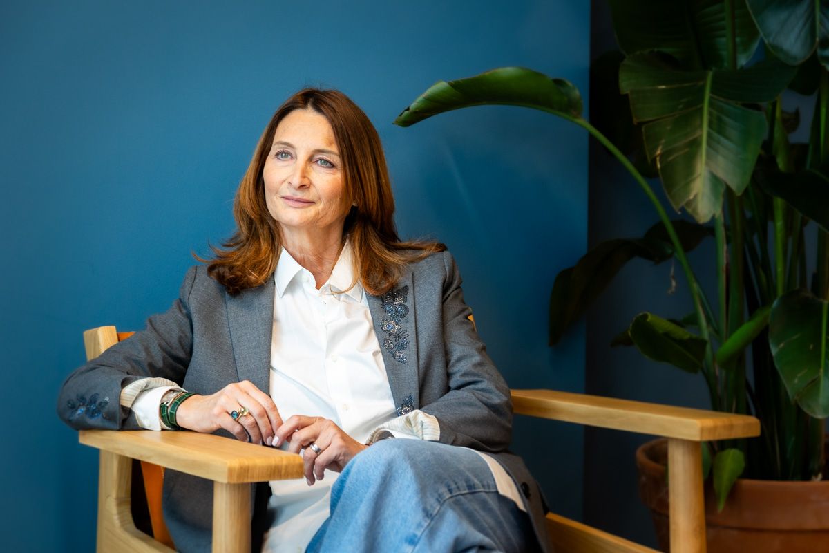 Professional wearing a grey blazer and white shirt sits at wooden desk with indoor plants in background.