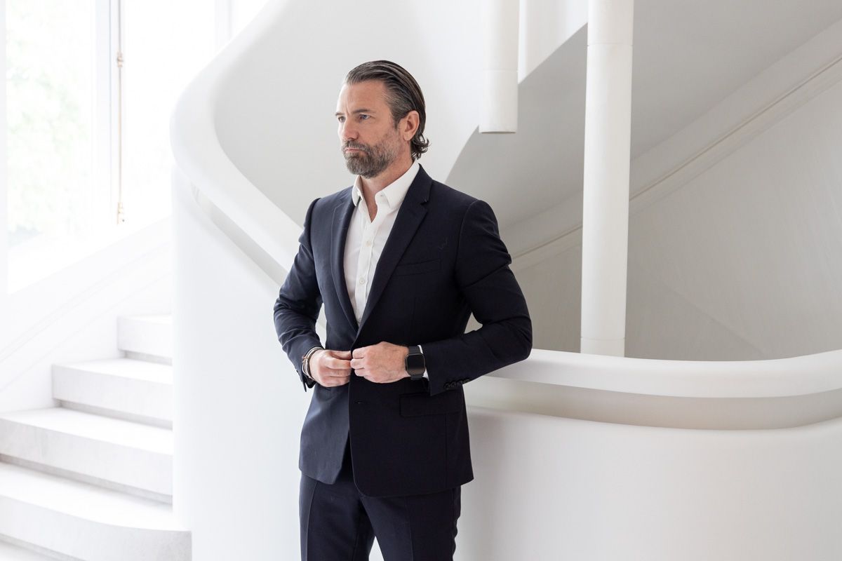 Professional businessman in a black suit looks at watch while standing in bright white modern office space.