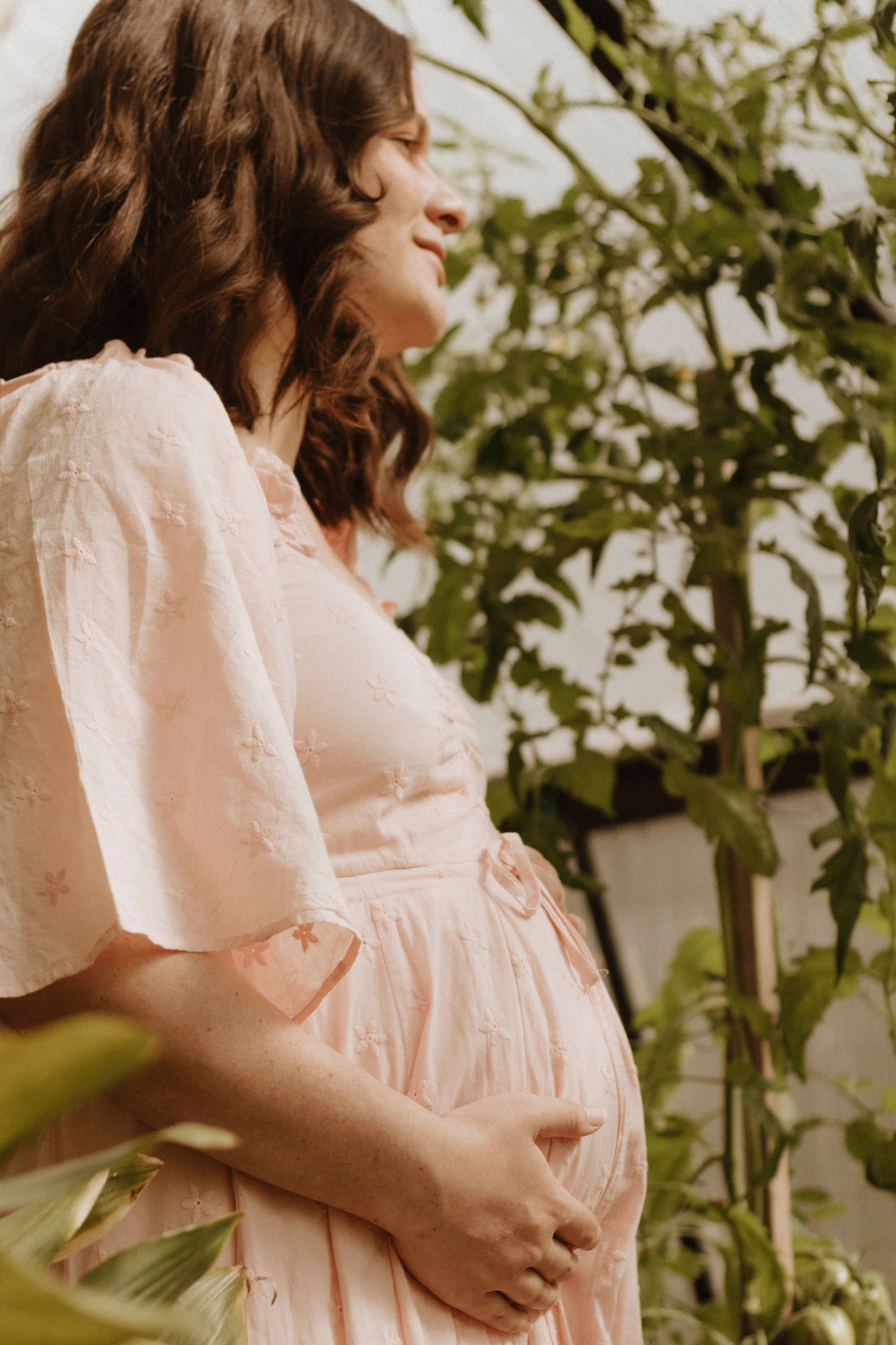 A pregnant woman in a flowing pink dress stands peacefully in a garden setting.