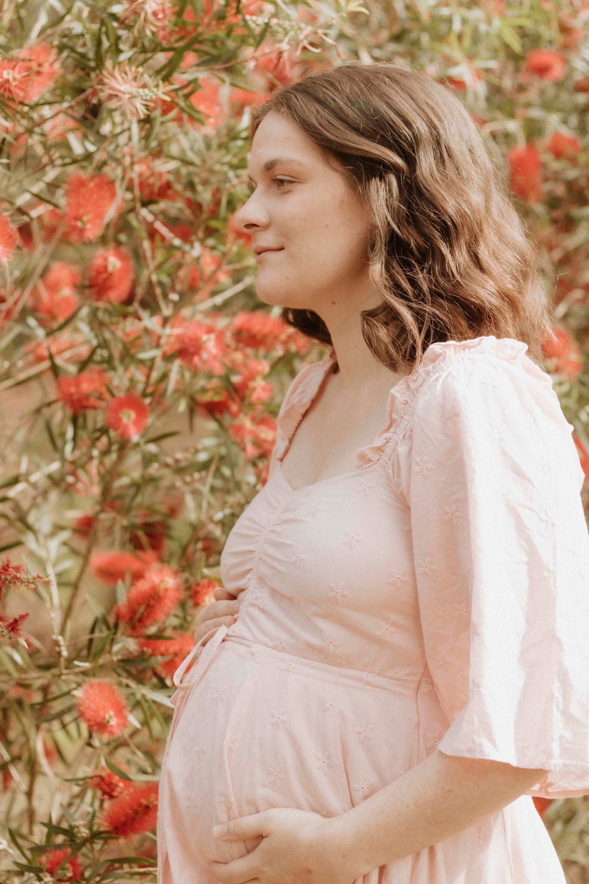 A pregnant woman in white dress poses among coral-colored flowers.