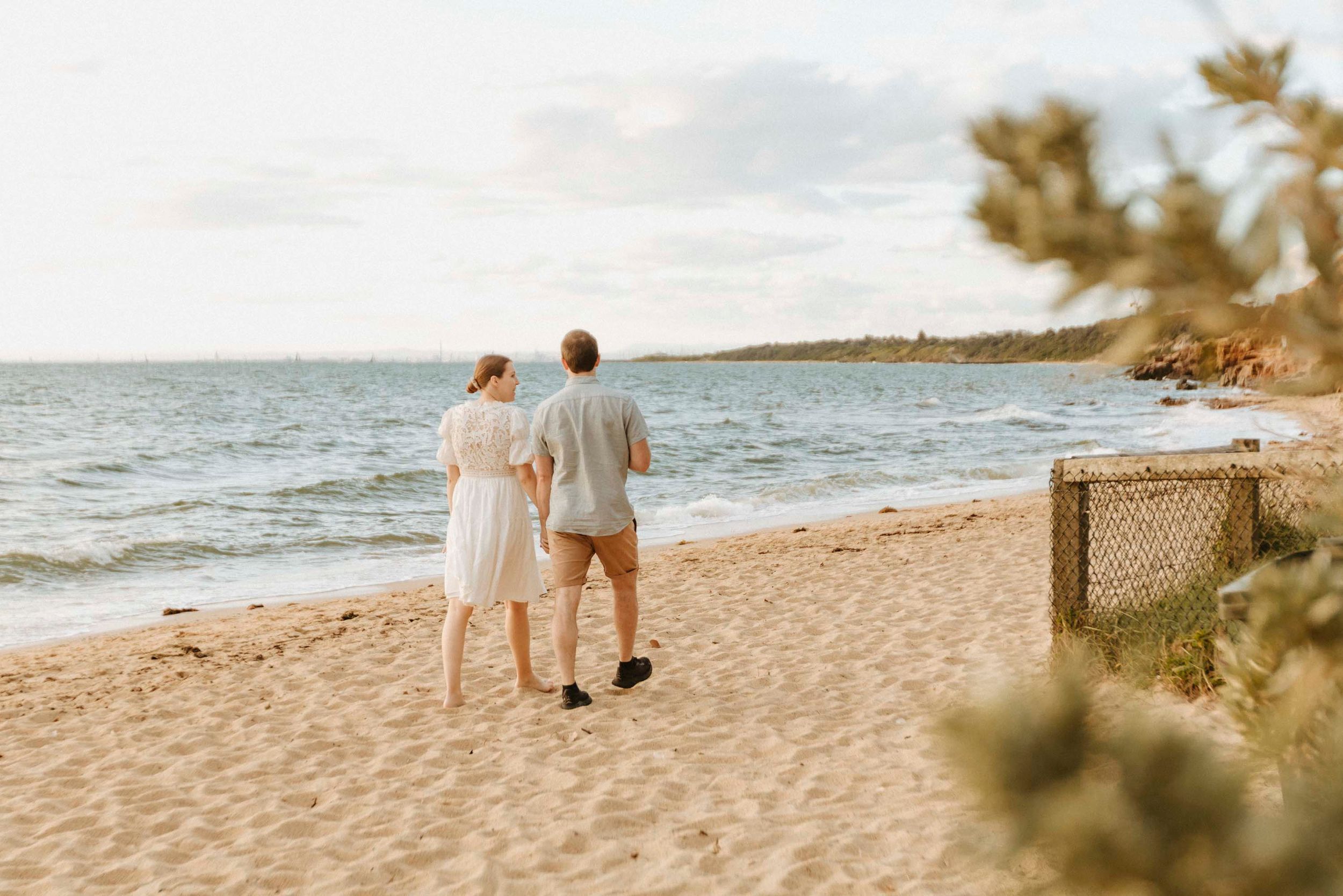 A couple walks hand in hand along a peaceful beach shoreline at sunset.