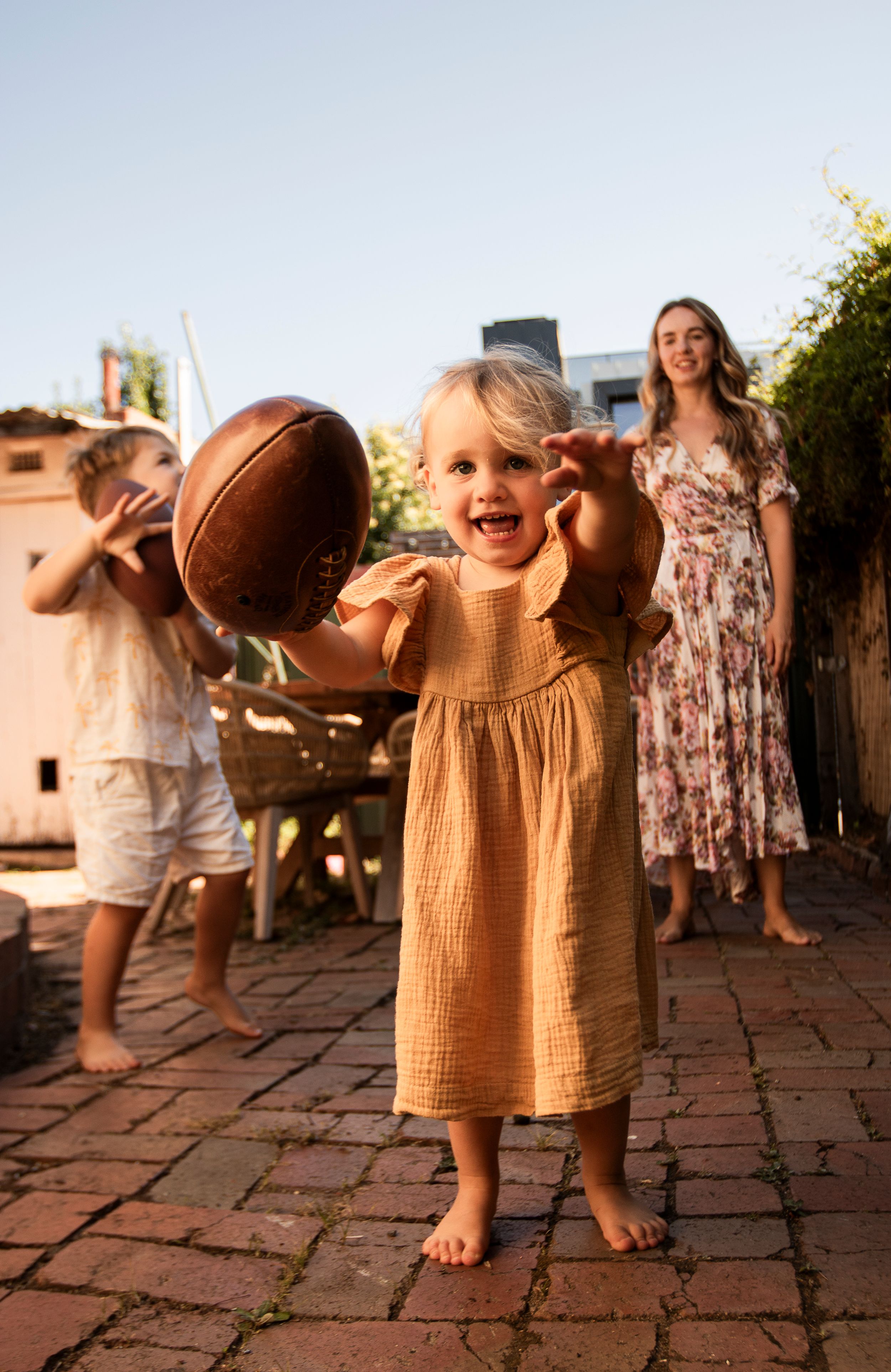 A toddler in mustard dress holds football while playing outdoors on brick patio.
