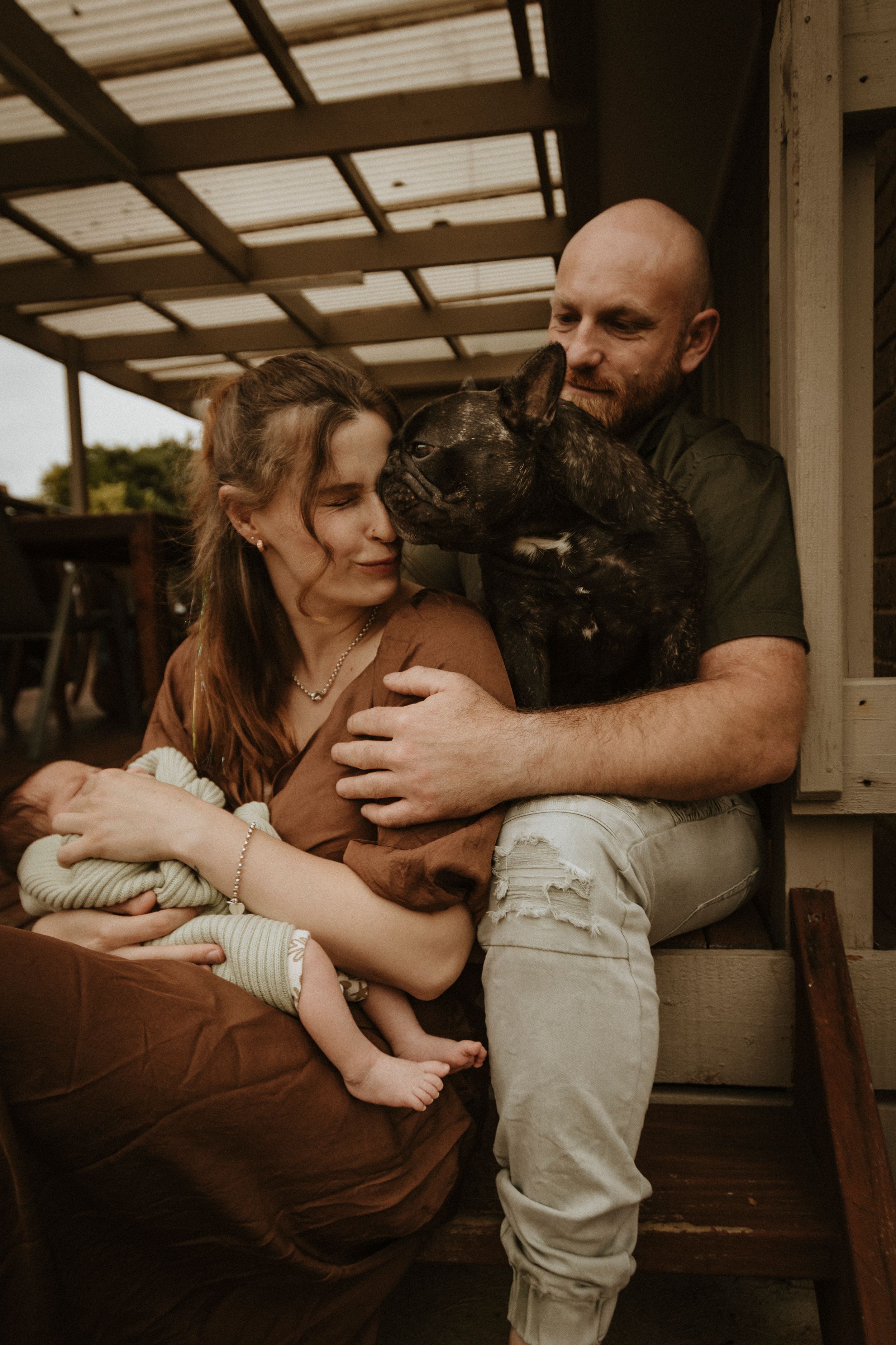 A family sitting on a porch with their French Bulldog and newborn baby.