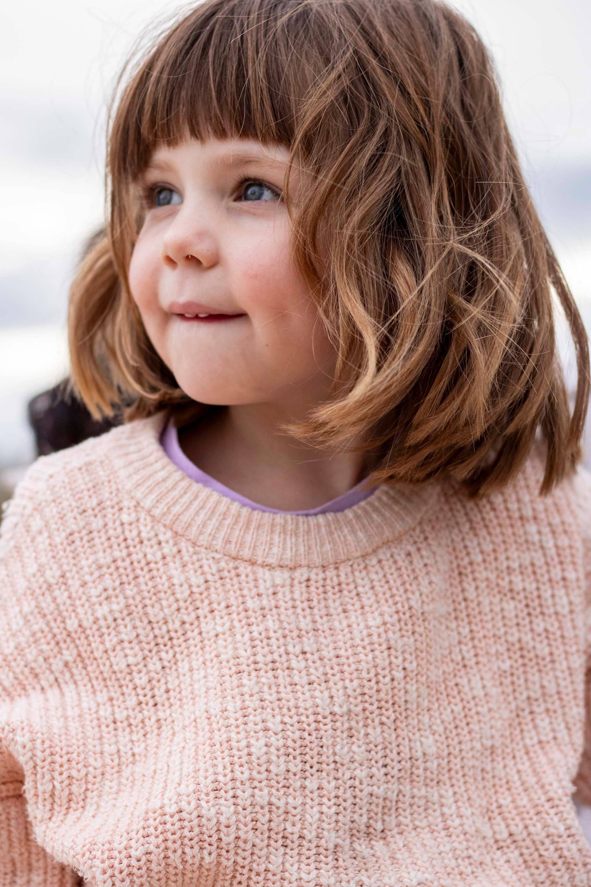 A child wearing a pink knit sweater smiles while looking off to the side.