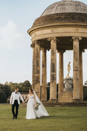 A wedding couple strolls hand in hand on manicured lawn near a classical domed temple structure with columns.