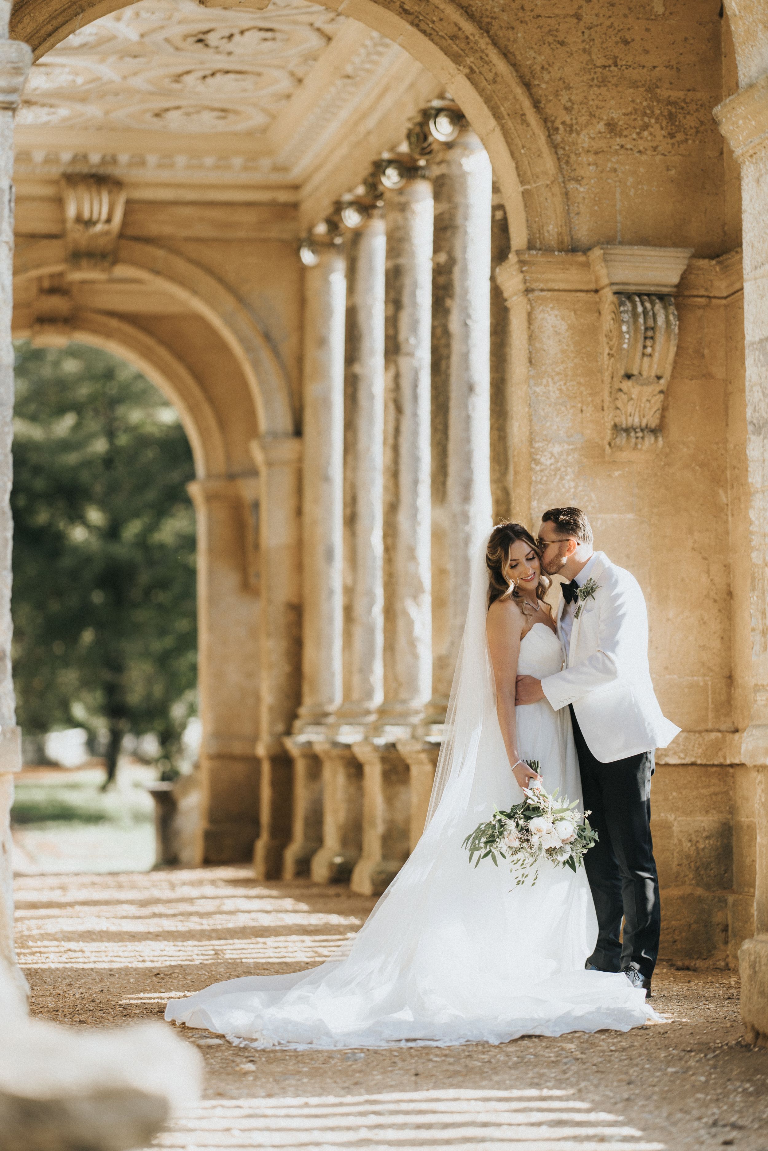Elegant wedding couple embraces under ornate stone archway with classical columns and sunlit garden backdrop.