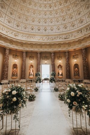 Elegant wedding ceremony setup in a circular room with ornate dome ceiling and floral arrangements lining the aisle.