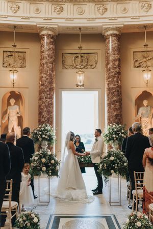 Wedding ceremony in a grand venue with marble columns and classical statues, decorated with white floral arrangements.