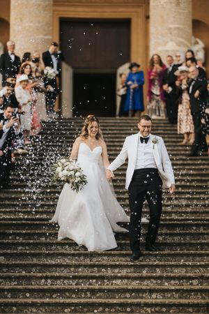 Newlyweds descend grand stone steps while guests shower them with confetti at elegant venue.